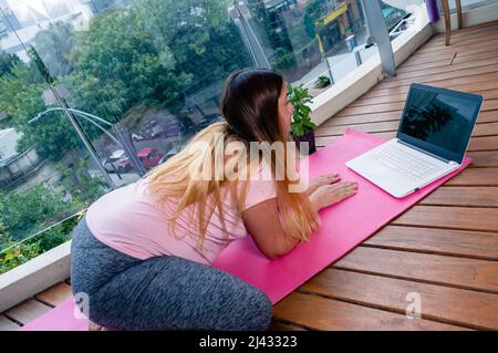 giovane donna caucasica incinta in classe online yoga con il suo laptop, sul balcone dell'appartamento facendo un asana su un tappeto rosa, guardando il portatile Foto Stock