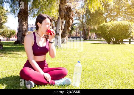 Una giovane atleta caucasica sta mangiando una mela rossa seduta sull'erba in un parco. Accanto a lei è una bottiglia di plastica trasparente di acqua. Indossa Foto Stock