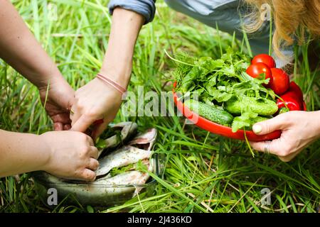 Donne senza volto che cucinano all'aperto in primavera, giorno d'estate. Le mani delle donne fanno roba di pesce fresco, tengono un piatto rosso di cetrioli lavati, pomodori, erbe. Persone Foto Stock