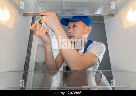 Un tecnico di sesso maschile in tute ripara un frigorifero rotto con un cacciavite. Concetto di riparazione frigorifero. Foto dall'interno del frigorifero Foto Stock