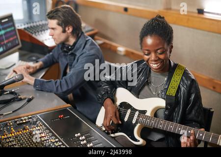Ritratto di una donna nera sorridente che suona la chitarra mentre produce musica in uno studio di registrazione professionale Foto Stock