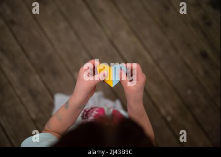 Vista dall'alto di un bambino che tiene carta a forma di cuore nei colori della bandiera ucraina - blu e giallo - in un'immagine concettuale degli orfani ucraini. Foto Stock