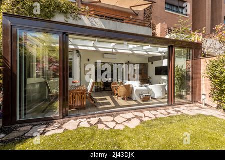 Sala da sole con ampie porte scorrevoli aperte con mobili in legno e vimini all'interno e una passerella in pietra e giardino erboso Foto Stock