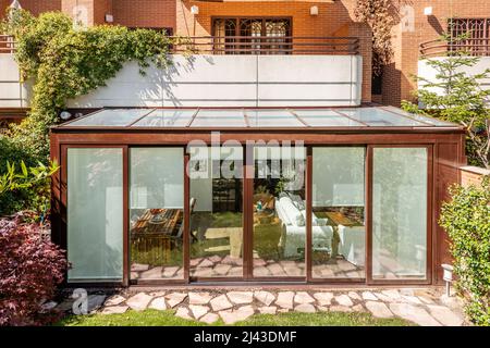 Terrazza coperta con struttura in alluminio marrone e vetro con accesso alla casa al piano terra e un giardino con un percorso di lastre di pietra e legge Foto Stock