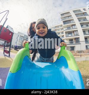 Felice ragazzo afroamericano con un cappello grigio di fronte a sua madre che sale su uno scivolo blu nel parco edifici sfondo vacchetta colpo. Foto di alta qualità Foto Stock