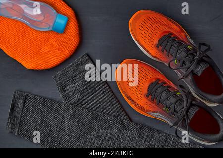 Sneakers arancioni, cappello in maglia arancione, leggings caldi per lo sport e una bottiglia d'acqua su un tavolo di cemento scuro. Attrezzatura per la corsa invernale Foto Stock