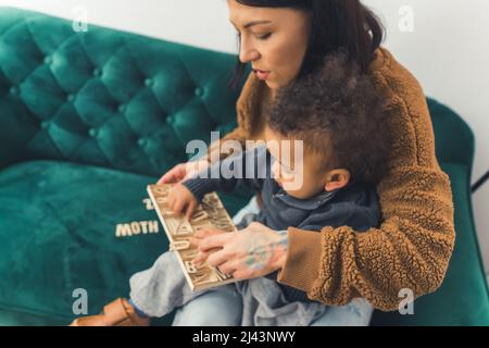 grazioso ragazzino arricciato seduto in grembo di sua madre che è seduto sul divano verde e guardando qualcosa con la madre medio pieno scatto spazio interno copia. Foto di alta qualità Foto Stock