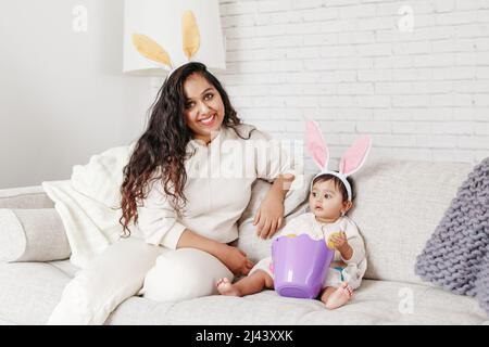 Madre indiana con bambina figlia in orecchie conigliere giocando uova giocattolo in cestino festeggiando la Pasqua. Foto Stock