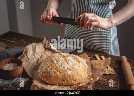 Un panettiere sta scattando foto di pane fresco fatto in casa sul suo telefono per un post sui social network. Il concetto di cottura dei prodotti da forno Foto Stock
