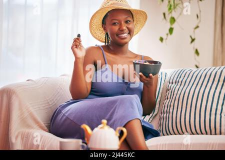 Sono a casa. Ritratto di una giovane bella donna che indossa un cappello da sole e che fa colazione sul divano a casa. Foto Stock