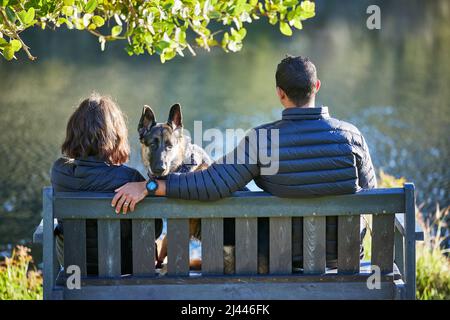 Non è un fine settimana lontano senza il nostro cane. Scatto retroguidato di una coppia seduta su una panca con il loro cane e guardando il lago. Foto Stock