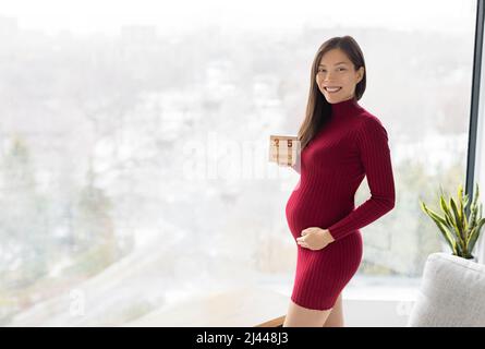 Donna incinta che mostra 25 settimane di gravidanza bump per maternità photoshoot tenendo blocchi di legno segno felice. Asiatico bello modello felice in piedi a casa Foto Stock