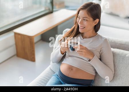 Donna incinta asiatica che beve una tazza di caffè a casa. Cibi e bevande di gravidanza da evitare Foto Stock