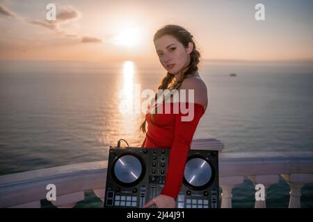 Le mani femminili del DJ suonano la musica mescolando e graffiando durante la festa serale con sfondo marino e caldo tramonto morbido. Primo piano di una console DJ controllata da Foto Stock