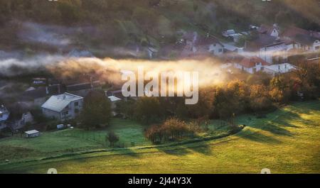 Campagna paesaggio rurale con villaggio sopra il fascio di sole in mattina nebbiosa. Foto Stock