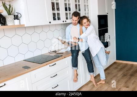 Una coppia di famiglie adorabili è in piedi in cucina a casa, facendo shopping online o chiacchierando con gli amici. Gioioso marito e moglie alla moda che usano un computer portatile Foto Stock