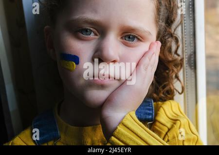Triste piccola ragazza Ucraina con bandiera di Ucraina sul suo viso seduto sul davanzale a casa. I bambini chiedono pace. Concetto di partecipazione di U Foto Stock