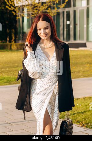 Elegante donna dai capelli rossi in abito bianco e giacca nera che cammina lungo la strada vicino all'edificio degli uffici Foto Stock