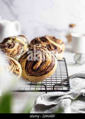 Panini alla cannella su rastrelliera con tovagliolo e fiori bianchi in primo piano. Composizione mattina di festa. Orientamento verticale Foto Stock