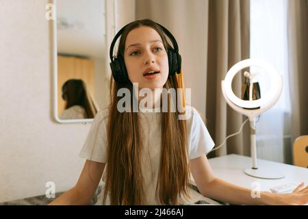 Bella ragazza caucasica adolescente, generazione Z con capelli lunghi in cuffie, a casa ascoltando musica audio e cantando insieme, sorridente. La vita reale, Foto Stock