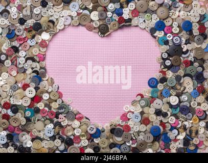 vari colori e bottoni di design disposti a forma di cuore su uno sfondo in tessuto rosa, presi dall'alto con spazio per la copia Foto Stock