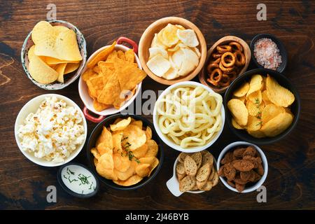 Cibo o spuntini malsani. Tutti i classici spuntini di patate con arachidi, anelli di popcorn e cipolle e pretzel salati in piatti da ciotola su vecchio sfondo di legno. Foto Stock