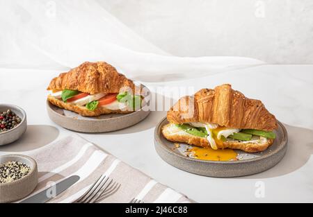 Gustosa prima colazione con croissant sandwich con uova in camicia, avocado, formaggio morbido, mozzarella e pomodoro in un piatto su sfondo bianco Foto Stock