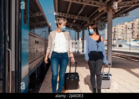 Due allegri amici femminili in maschere protettive camminando e chiacchierando sulla piattaforma alla stazione ferroviaria Foto Stock