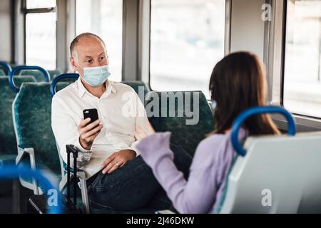 Un paio di amici che indossano la maschera e parlano bene mentre si viaggia in treno Foto Stock