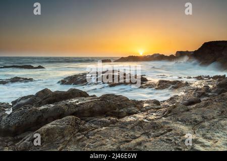 Tramonto sulla spiaggia di Leo Carrillo, vicino a Malibu, California. Sole arancione appena sopra l'orizzonte. Spiaggia rocciosa e acqua setosa e fluente. Scogliere in lontananza. Foto Stock