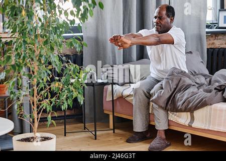 Uomo anziano attivo in t-shirt di cotone bianco e pantaloni grigi che mantengono le braccia tese davanti a se stesso mentre si esercita a letto dopo il sonno Foto Stock