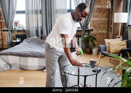 Uomo anziano contemporaneo in casa spolverare piccolo tavolo rotondo di vetro con detergente in soggiorno durante le attività domestiche Foto Stock