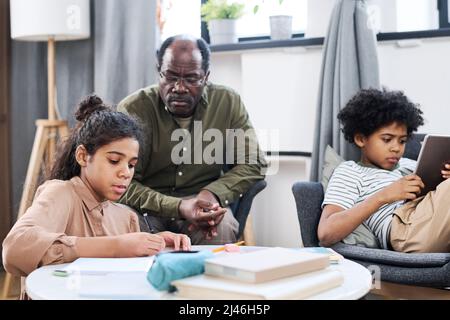 Scolgirl youthful che fa le note nel copybook contro il suo nonno che si siede in poltrona e fratello usando il tablet mentre fa i compiti Foto Stock