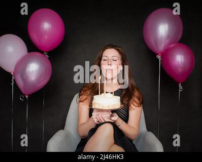 Donna graziosa che soffia le candele fuori sulla torta di compleanno con i palloncini. Foto Stock