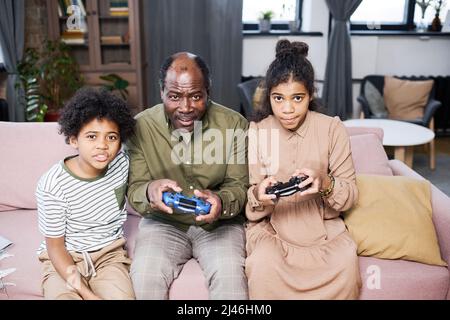 Due simpatici scolari multietnici e il loro nonno giocano a giocare a tempo libero davanti al televisore mentre si siedono sul divano in soggiorno Foto Stock