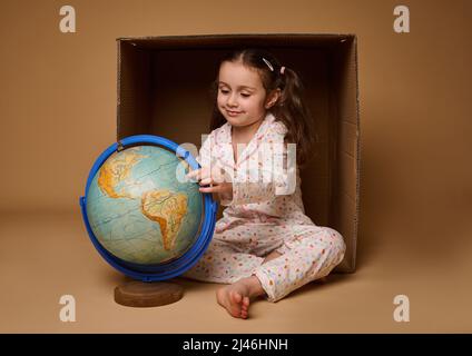 Adorabile bambina in pigiama seduto davanti a una scatola di cartone ed esaminando il globo terrestre, isolato su sfondo beige con spazio copia. Amore Foto Stock