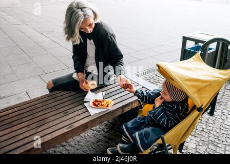 La donna si siede sulla panchina accanto al passeggino con il figlio e mangia curry wurst Foto Stock