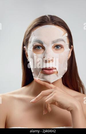 Trattamenti viso e bellezza. Donna con un panno maschera idratante sul viso su sfondo grigio. Procedura cosmetica. Foto Stock