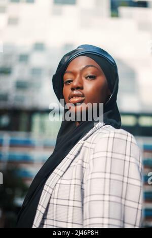 Vista laterale basso angolo di sicurezza afro-americana musulmana donna vestito in elegante scacchi blazer con foulard Islamico e berretto in pelle guardando la macchina fotografica con fiducia Foto Stock