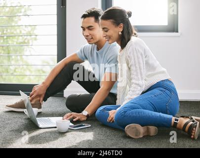 Volevo mostrarti i mobili che ho appena ordinato. Scatto di una giovane coppia seduta sul pavimento insieme nella nuova casa e utilizzando un computer portatile. Foto Stock