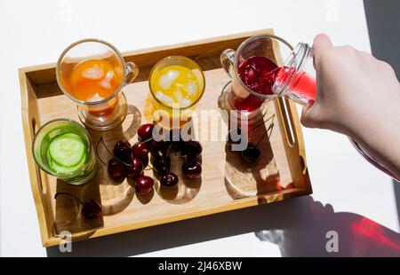 Un vassoio di legno con bevande colorate: Limonata arancione, gialla e cetriolo. Nelle vicinanze si trovano le ciliegie. Una mano versa il succo rosso da un decanter in un bicchiere. C Foto Stock