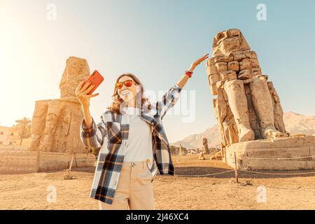 Travel blogger ragazza scatta foto selfie su uno smartphone presso i famosi due Colossi di Memnon - enormi statue in rovina del faraone Amenhotep III L Foto Stock