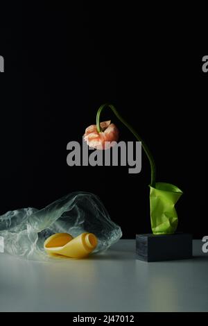 Verticale concettuale STILL LIFE composizione di fiori in tazza di plastica sbriciolata, busta di plastica e bottiglia sul tavolo, sfondo nero parete Foto Stock