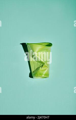 Verticale dalla vista dall'alto scatto di una tazza di plastica verde sminuzzata su sfondo verde azzurro chiaro, consumo consapevole e concetto di riciclaggio Foto Stock