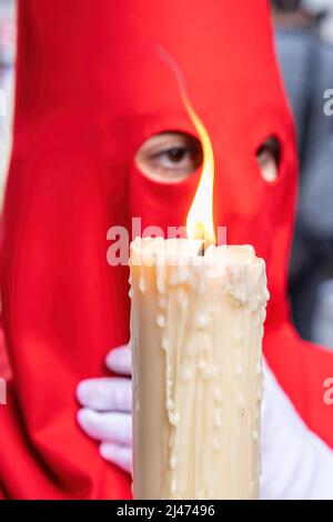 La candela in fiamma di un giovane Nazareno con un cappuccio rosso che fa la sua stazione di penitenza nella processione della settimana Santa. Fuoco selettivo solo con fiamma candela Foto Stock