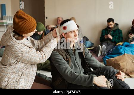 Ritratto di giovane donna bandaging capo di giovane ferito in rifugio rifugiati, spazio copia Foto Stock