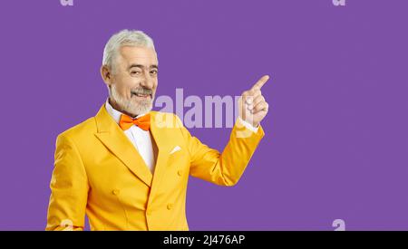 L'uomo anziano felice in abito giallo su sfondo viola sorride e punta il dito sullo spazio della copia Foto Stock