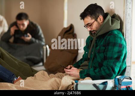 Ritratto laterale del giovane uomo nascosto in rifugio durante la guerra o la crisi e tenendo smartphone Foto Stock