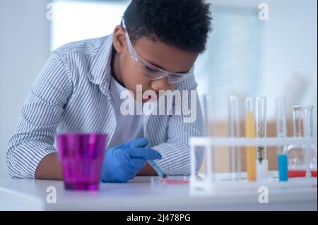 Un ragazzo dai capelli scuri in un camice da laboratorio che tiene una siringa con un reagente e che ha un aspetto occupato Foto Stock