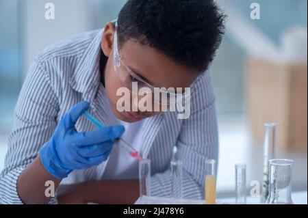 Un ragazzo dai capelli scuri in un camice da laboratorio che tiene una siringa con un reagente e che ha un aspetto occupato Foto Stock